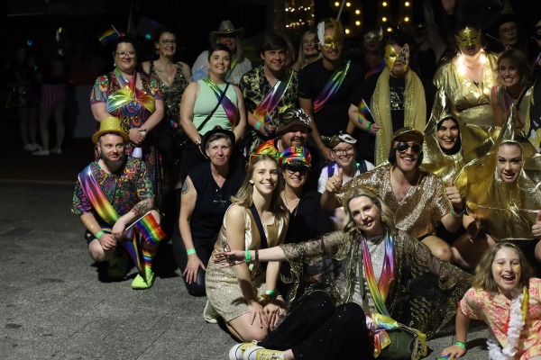 A group of people gathered together at night wearing costumes that have various rainbow lgbtqia+ patterns on them