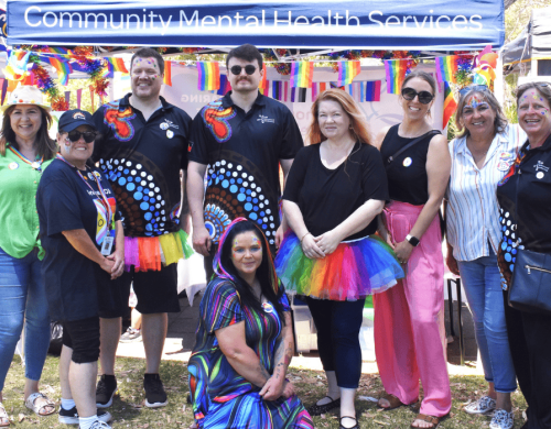Nine people standing together in rainbow outfits and face paint