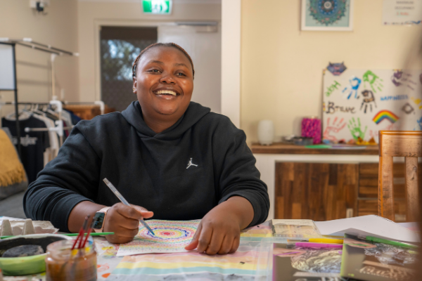 A woman sitting at a table and smiling at the camera while she draws a brightly coloured pattern