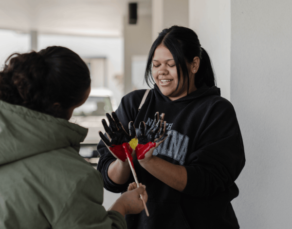 A woman smiling while another perrson paints the patterns of the aboriginal flag red, black and yellow on her hands