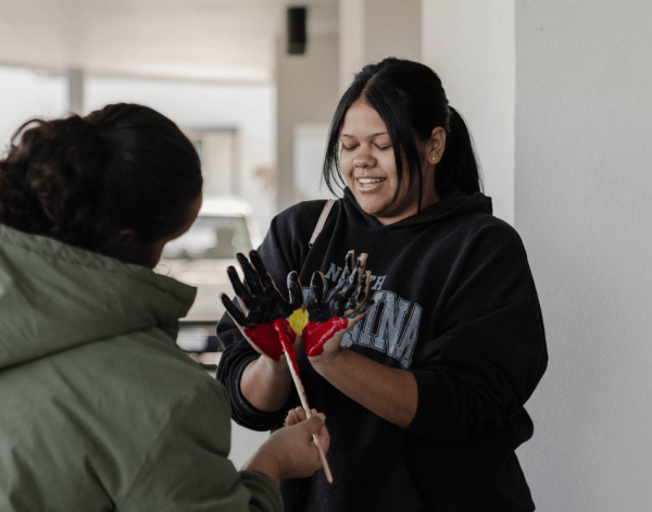 A woman smiling while another perrson paints the patterns of the aboriginal flag red, black and yellow on her hands