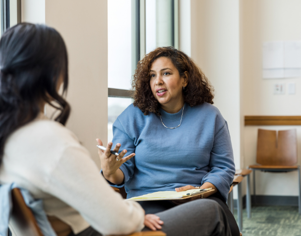 A woman in a blue top explaining something to another woman while holding a clipboard and gesticulating