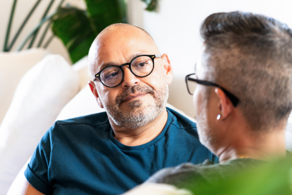 A man with glasses and a blue shirt sitting on a couch and listening to another man