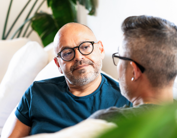 A man with glasses and a blue shirt sitting on a couch and listening to another man