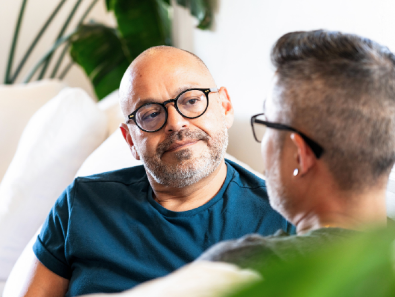 A man with glasses and a blue shirt sitting on a couch and listening to another man
