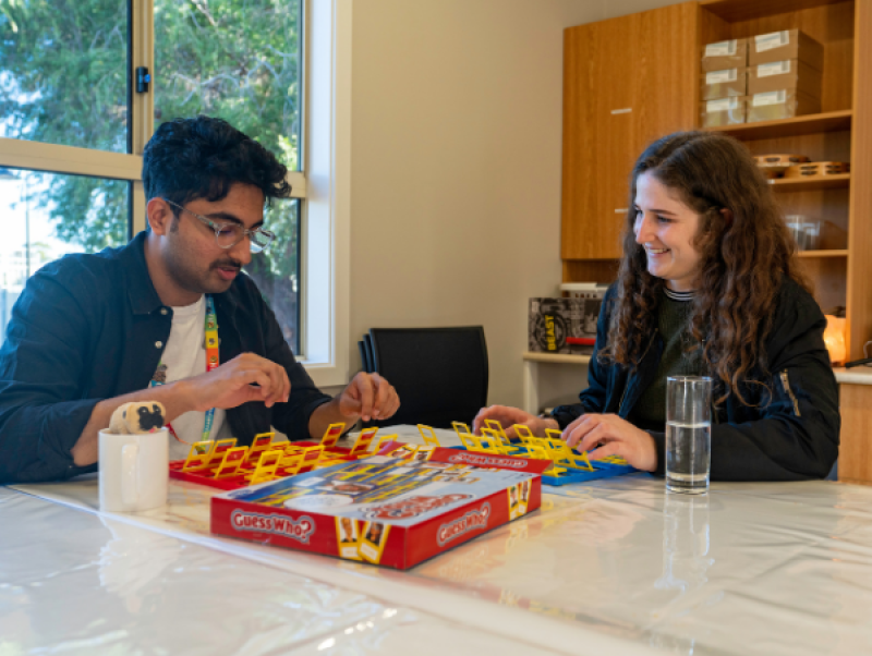 A man and a woman sitting at a table indoors and playing guess who