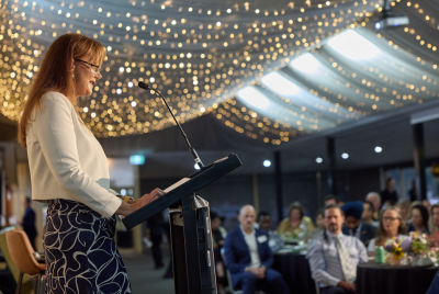 A woman with glasses standing at a podium and giving a presentation to a room full of people with a lot of lights on the ceiling