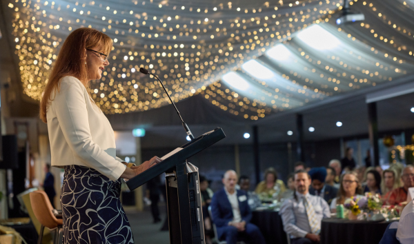 A woman with glasses standing at a podium and giving a presentation to a room full of people with a lot of lights on the ceiling