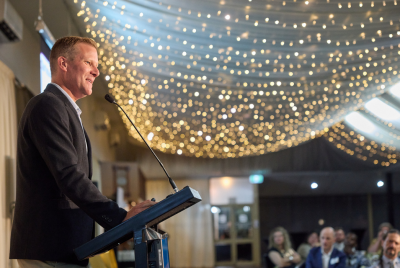 A man in a suit giving a speech at a podium to a room full of people with many lights hanging from the ceiling