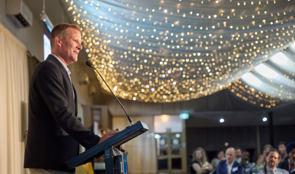A man in a suit giving a speech at a podium to a room full of people with many lights hanging from the ceiling