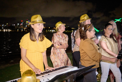 A group of women in singing in yellow fedoras, while one of them is playing the keyboard