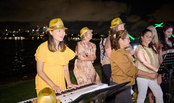 A group of women in singing in yellow fedoras, while one of them is playing the keyboard