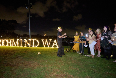 A group of people in yellow fedoras singing with one woman playing a keyword in front of a Richmind WA light display