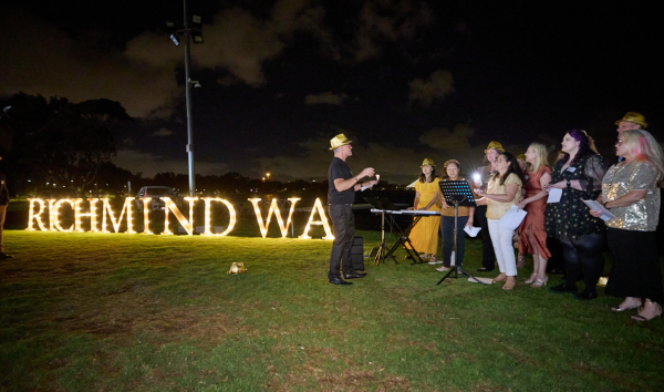 A group of people in yellow fedoras singing with one woman playing a keyword in front of a Richmind WA light display
