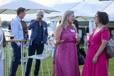 Two women speaking at at event on the grass while two men drink from cups in the background