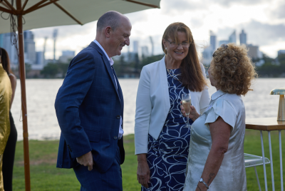 Two women and a man smiling and talking to each other at an event while standing on the grass in front of the river