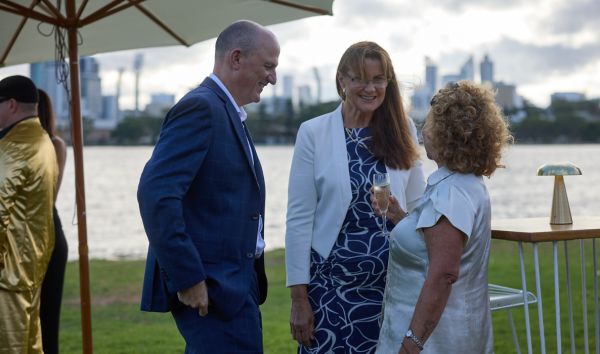 Two women and a man smiling and talking to each other at an event while standing on the grass in front of the river