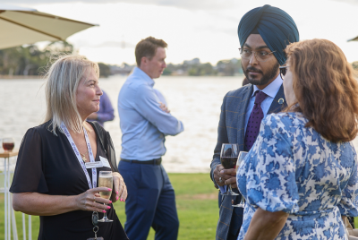 A man and two women standing together and speaking at an event on the grass in front of the river while a man walks past in the background