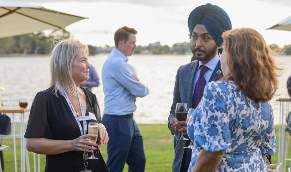 A man and two women standing together and speaking at an event on the grass in front of the river while a man walks past in the background