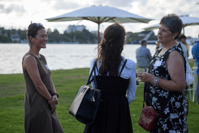 Three women standing together and talking at an event on the grass, in front of the river