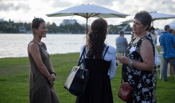 Three women standing together and talking at an event on the grass, in front of the river
