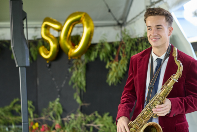 A man in a red suit jacket playing saxophone on a stage with gold balloons of the number 50 in the background