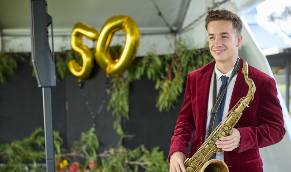 A man in a red suit jacket playing saxophone on a stage with gold balloons of the number 50 in the background