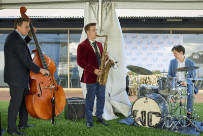 A band playing together at an event with a saxophonist in a red suit jacket, a double bass player in a black suit and a a drummer in a blue suit