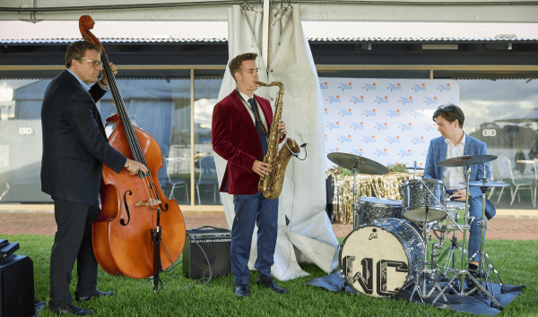 A band playing together at an event with a saxophonist in a red suit jacket, a double bass player in a black suit and a a drummer in a blue suit