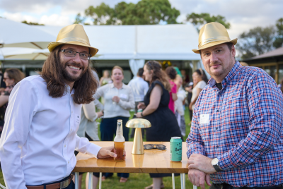 Two men sitting at a table with drinks and staring in the camera while wearing gold fedoras