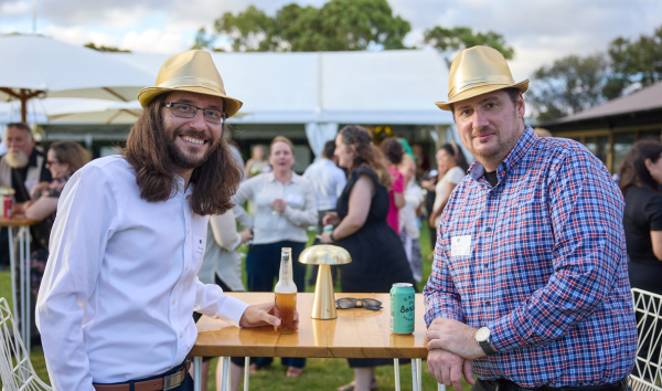 Two men sitting at a table with drinks and staring in the camera while wearing gold fedoras