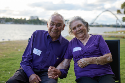 An older man and an older woman in purple shirts and sitting in black chairs on the grass and holding hands