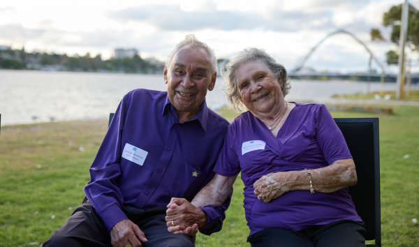 An older man and an older woman in purple shirts and sitting in black chairs on the grass and holding hands