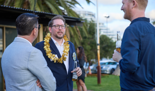 Three men in suits holding drinks while talking at an event while one of them wears a gold necklace