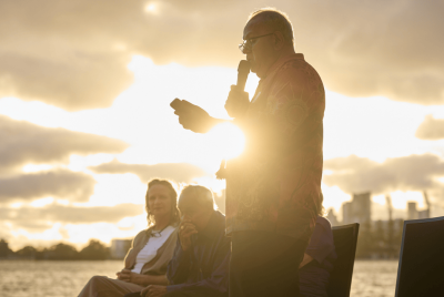 A man giving a speech into a microphone while the sun is setting and few people sit in chair behind him