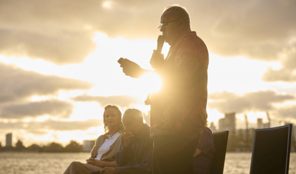 A man giving a speech into a microphone while the sun is setting and few people sit in chair behind him