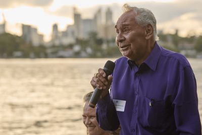 A man in a purple shirt giving a speech into a microphone while standing in front of the river
