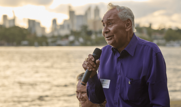 A man in a purple shirt giving a speech into a microphone while standing in front of the river