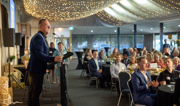 A man in a blue suit standing on a stage and giving a speech into a microphone to a group of people at an event