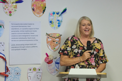 Woman giving a speech at a podium with decorated paper masks on the wall behind her