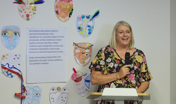 Woman giving a speech at a podium with decorated paper masks on the wall behind her