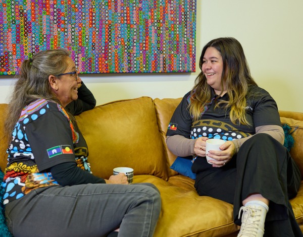 Two women smiling and chatting together on a couch while holding mugs