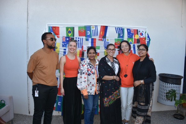 A group of six people smiling and posing for a photo in front of a backdrop with many different country flags