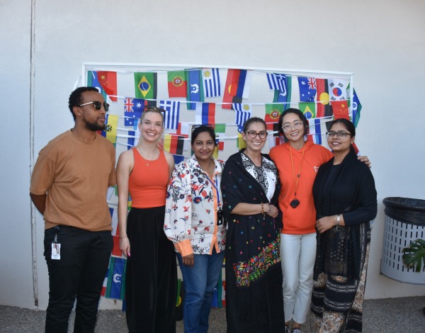 A group of six people smiling and posing for a photo in front of a backdrop with many different country flags
